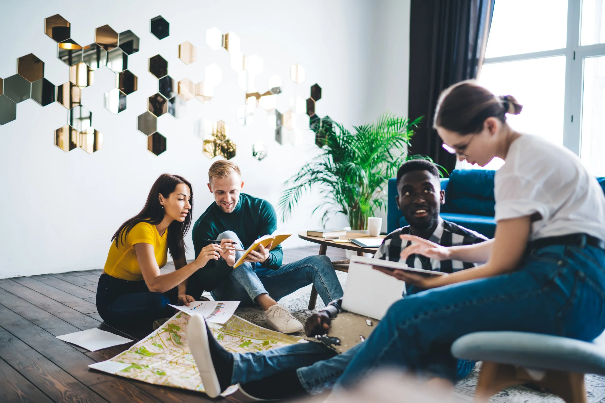 A group of four diverse adults sitting on the floor in a modern room, discussing and reviewing maps and documents. The room features geometric wall decor and indoor plants, with natural light coming from a large window.