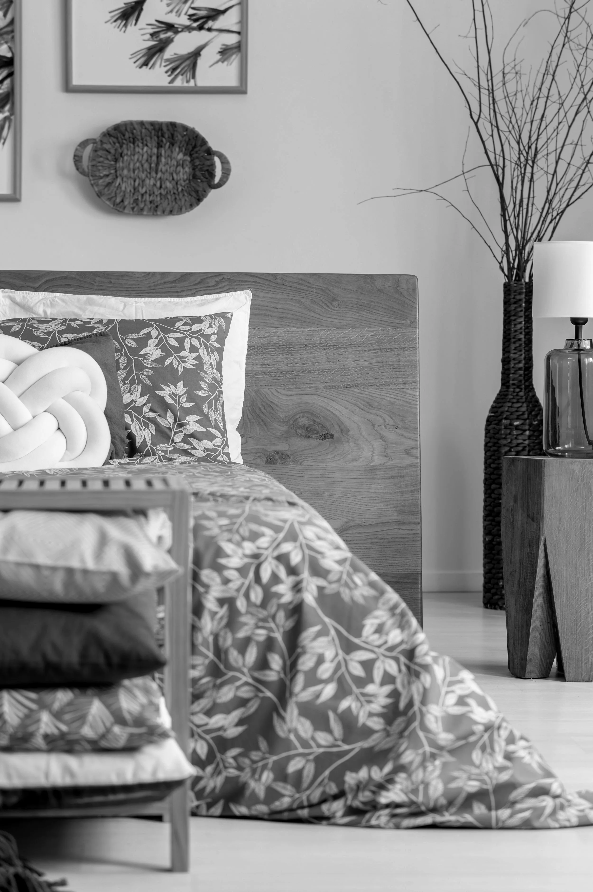 Modern bedroom with wooden headboard, patterned bedding, decorative pillows, and a woven basket on the wall. A side table holds a lamp and a wicker vase with branches.