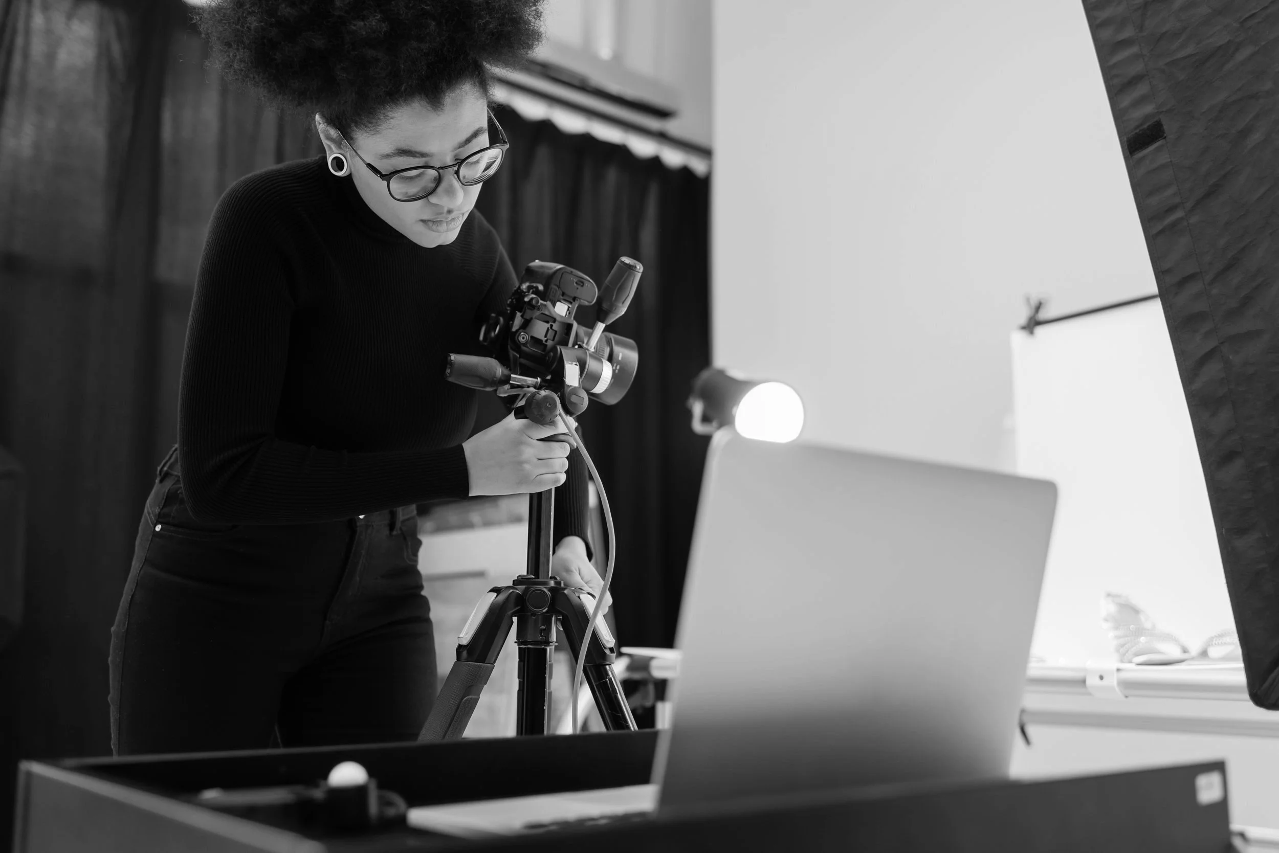 Person adjusting camera on tripod in a photography studio setup with laptop and lights.