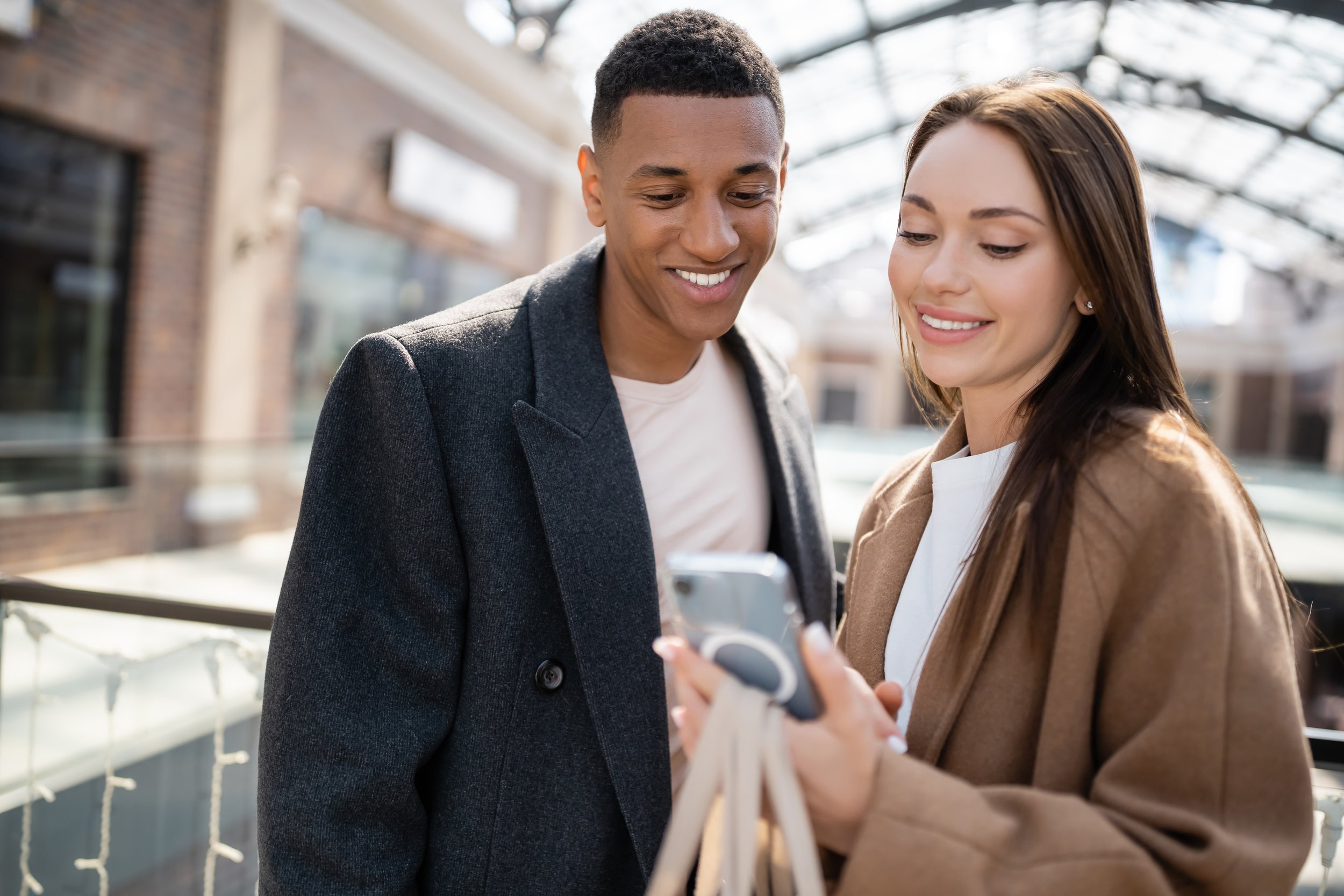 Smiling couple looking at a smartphone indoors, wearing coats.