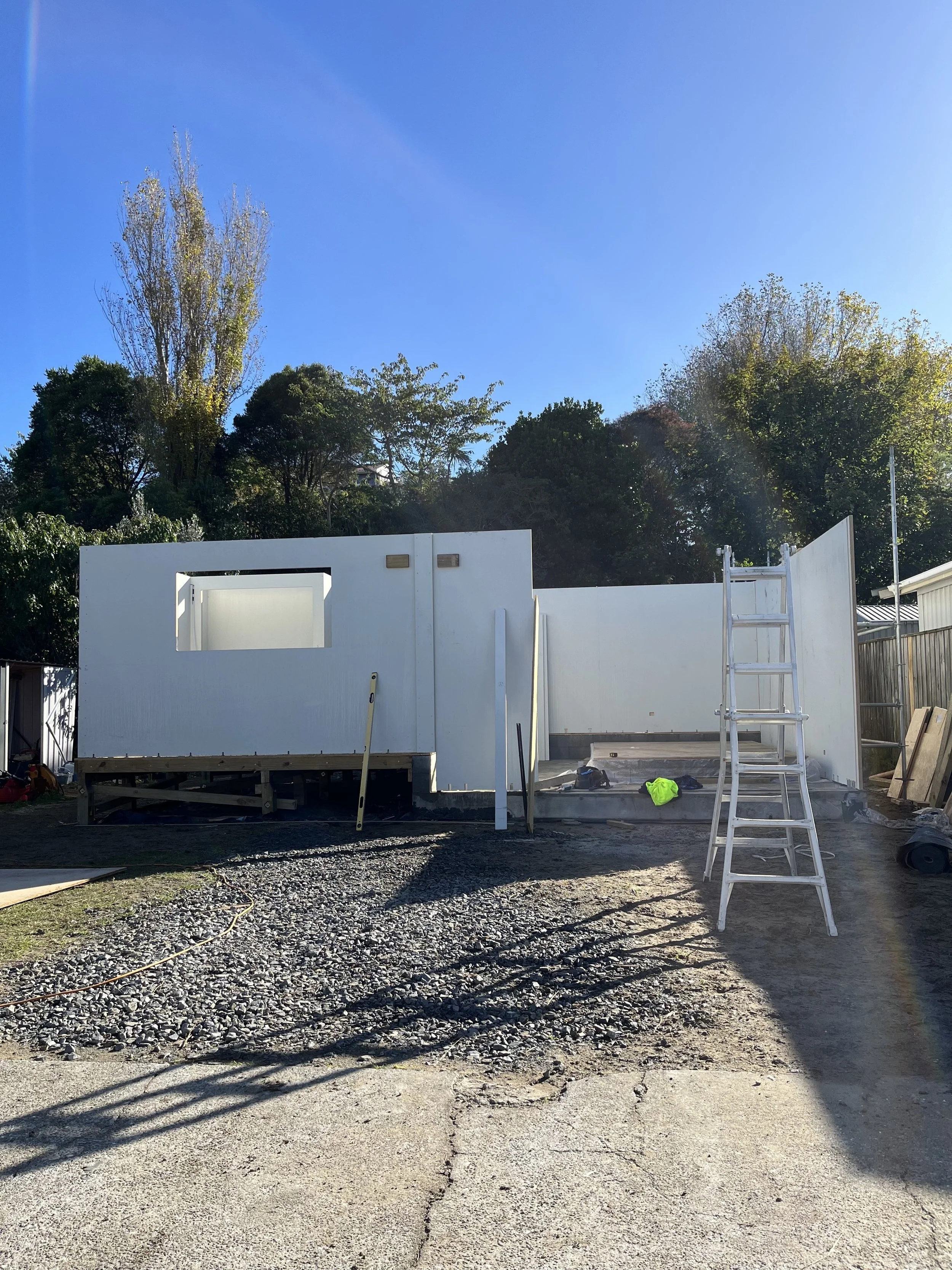 Construction site with white prefabricated wall sections, a ladder, and construction materials on a gravel surface under a blue sky, with trees in the background.