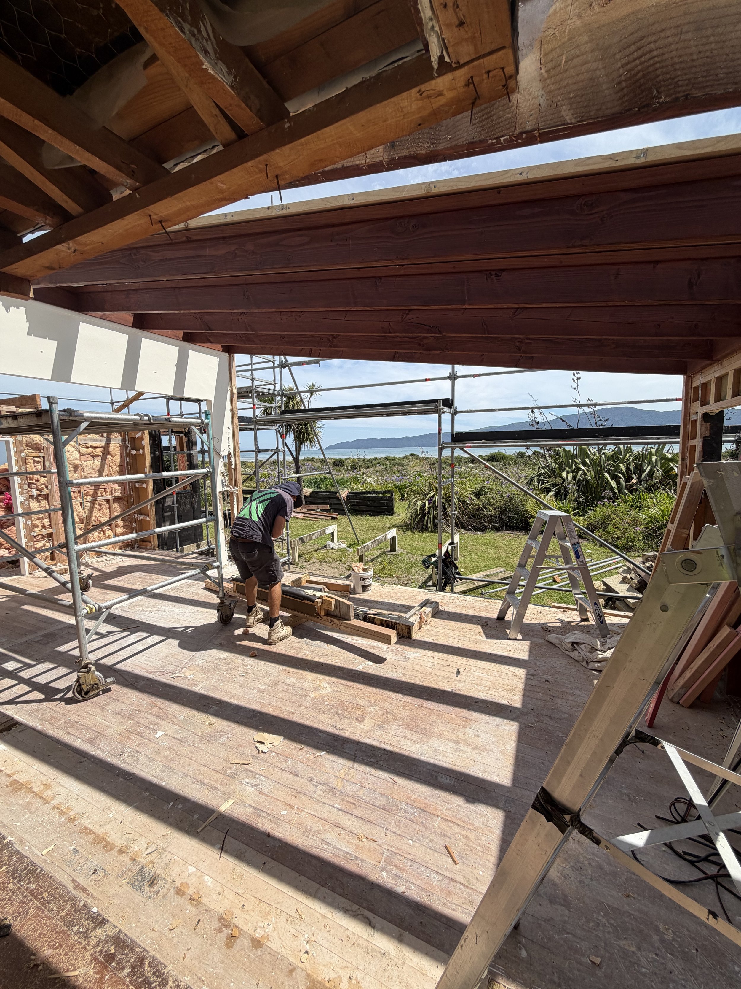 A person working on a construction site on a wooden deck outdoors, with scaffolding and ladders, overlooking a grassy area with trees and mountains in the background.