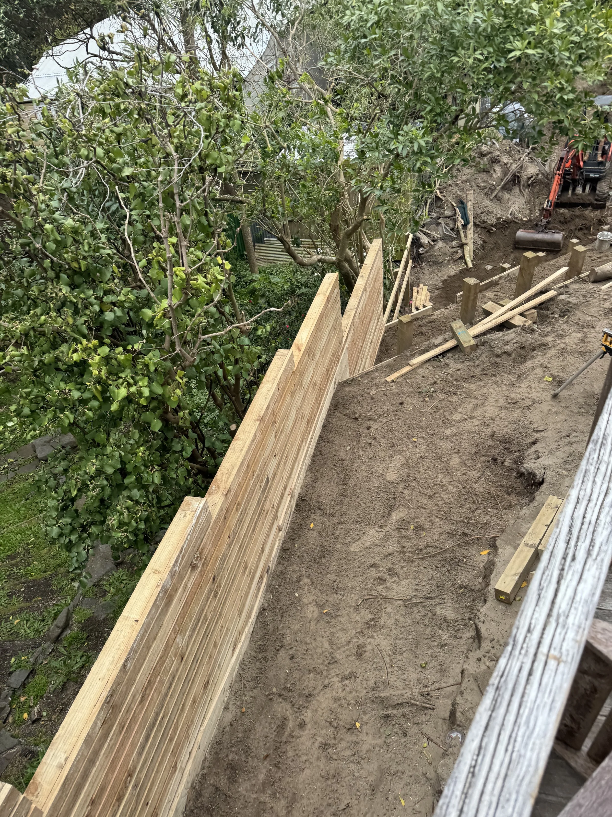 Construction site with partially built wooden fence, dirt ground, construction tools, and an excavator in the background.