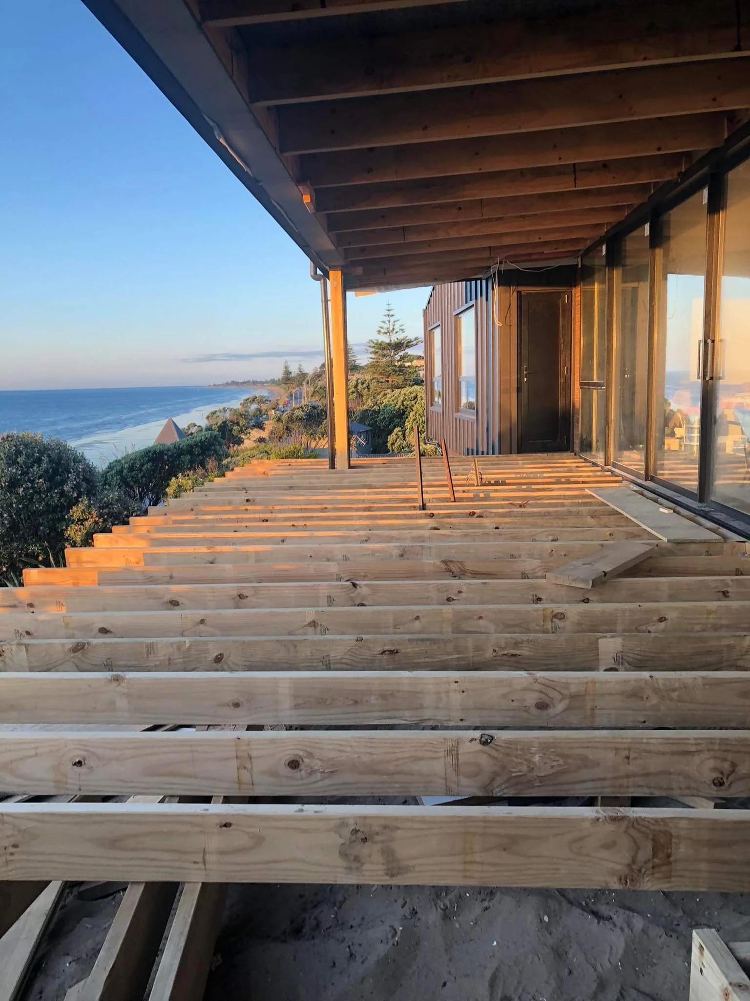 Construction of a balcony or deck with wooden beams and flooring overlooking the ocean, sunset lighting.
