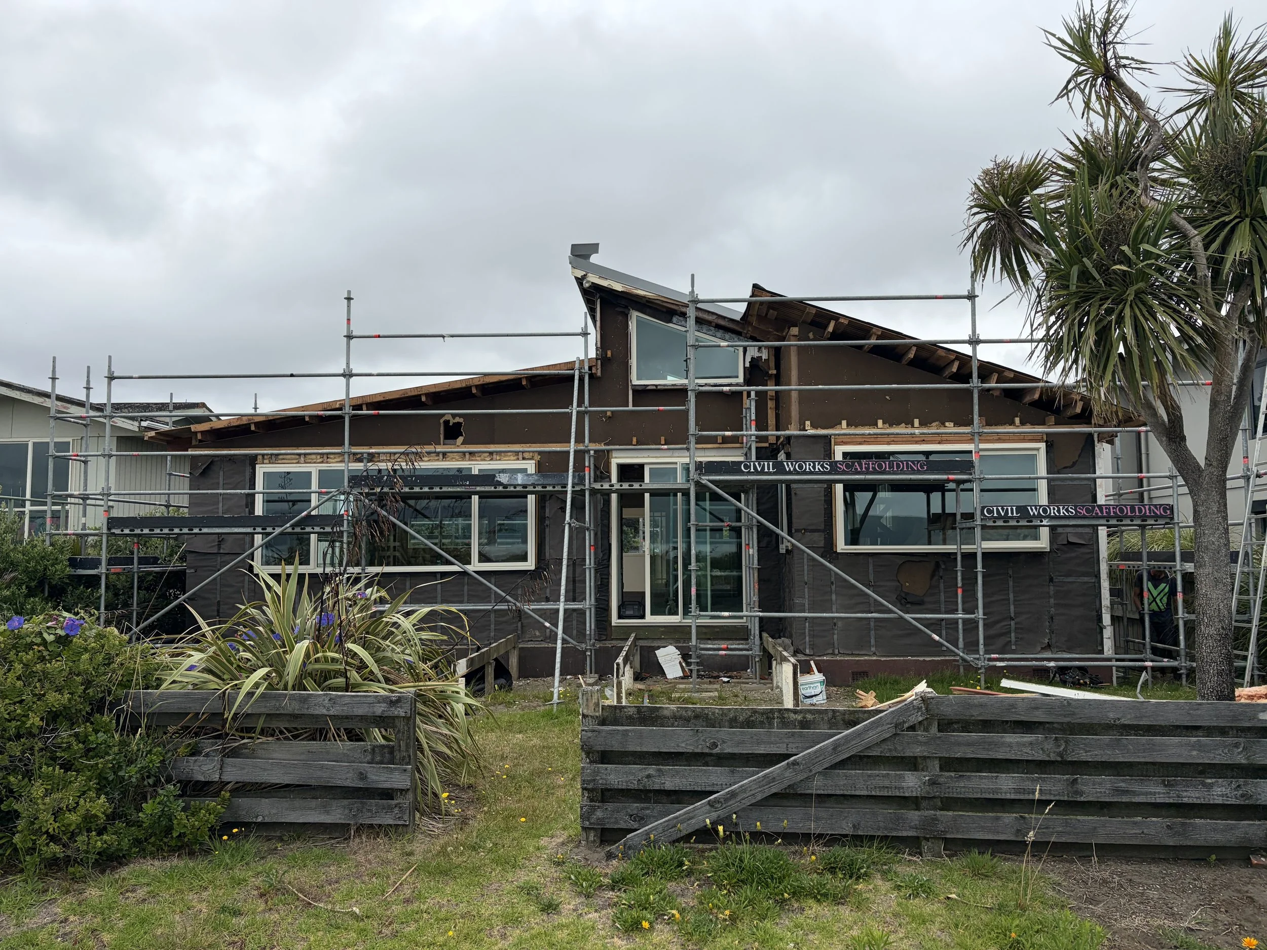 House under construction with scaffolding, dark exterior walls, large windows, and a sloped roof on a cloudy day.