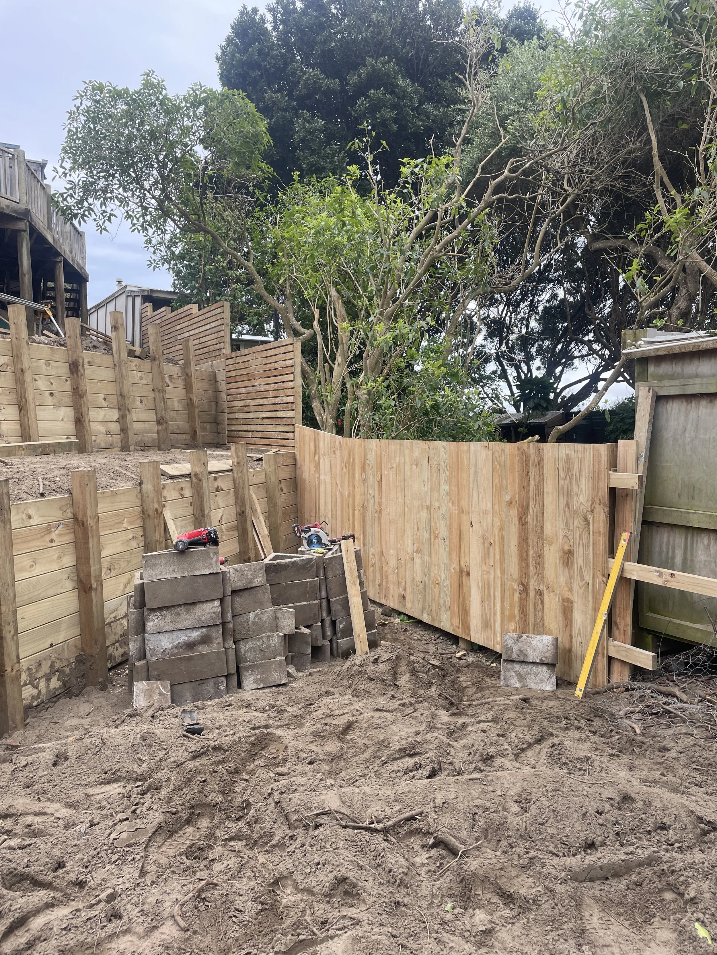 A backyard in the process of fence installation featuring stacks of concrete blocks, tools, and newly built wooden fence sections surrounded by trees and sky.