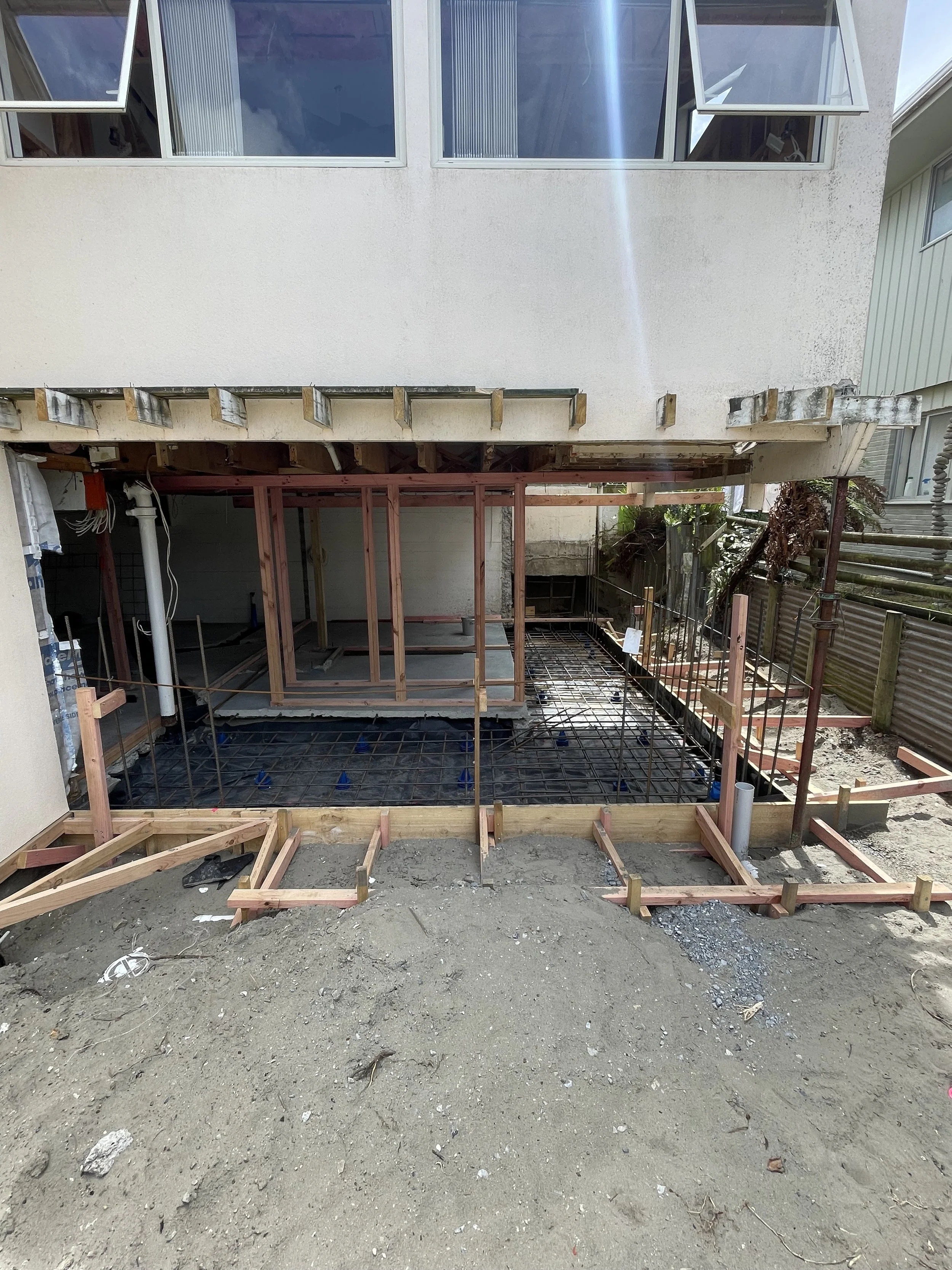 Construction site with wood framing and rebar for a new patio or extension under a house, with dirt in the foreground and existing building above.