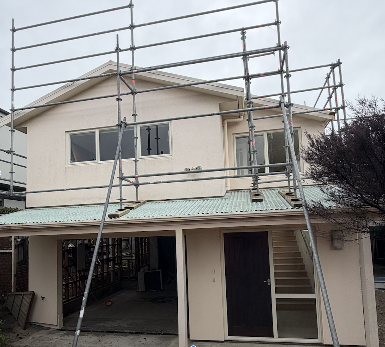 A house under construction with scaffolding around it, showing the upper floor and exterior walls, stairs, and garage area.