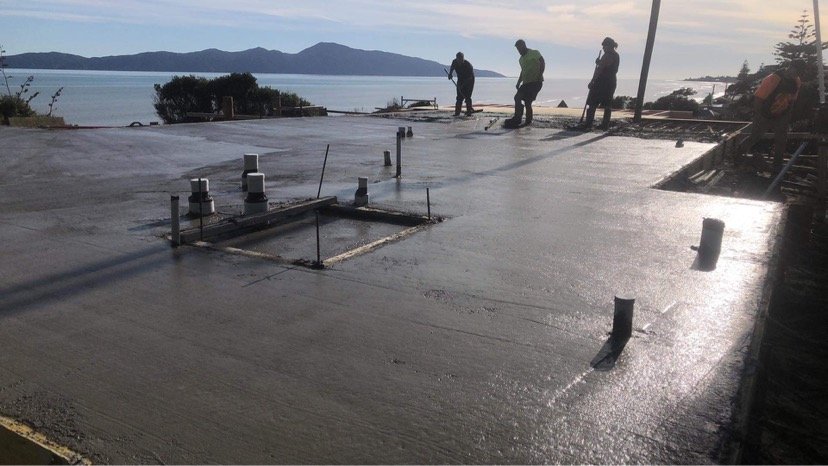 Construction workers pouring concrete on a building foundation with a scenic view of mountains and water in the background.