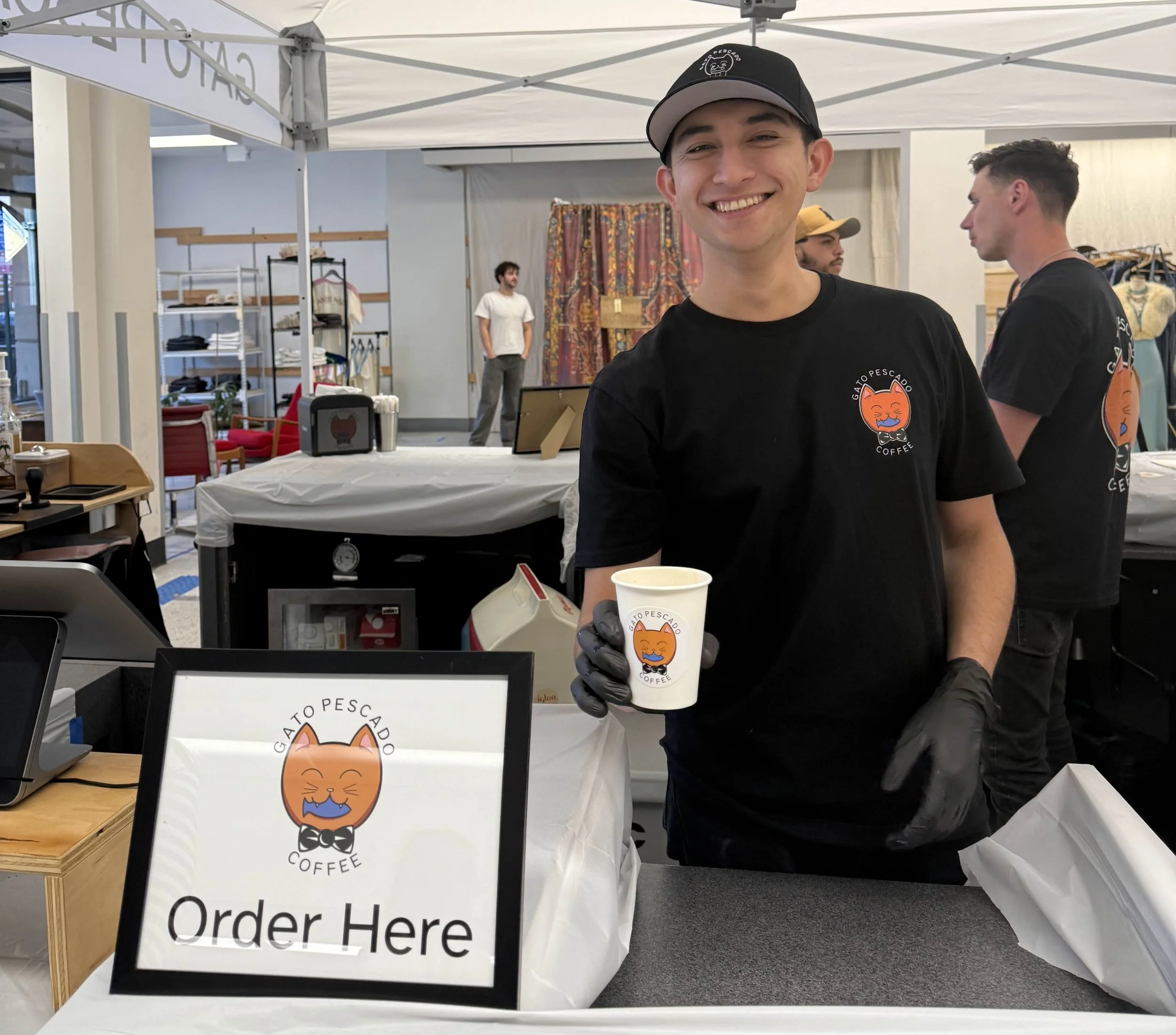 Smiling young man wearing a black cap and black t-shirt with a logo, holding a paper coffee cup with the same logo, at a coffee booth. The sign on the table reads 'Order Here' with the same logo.