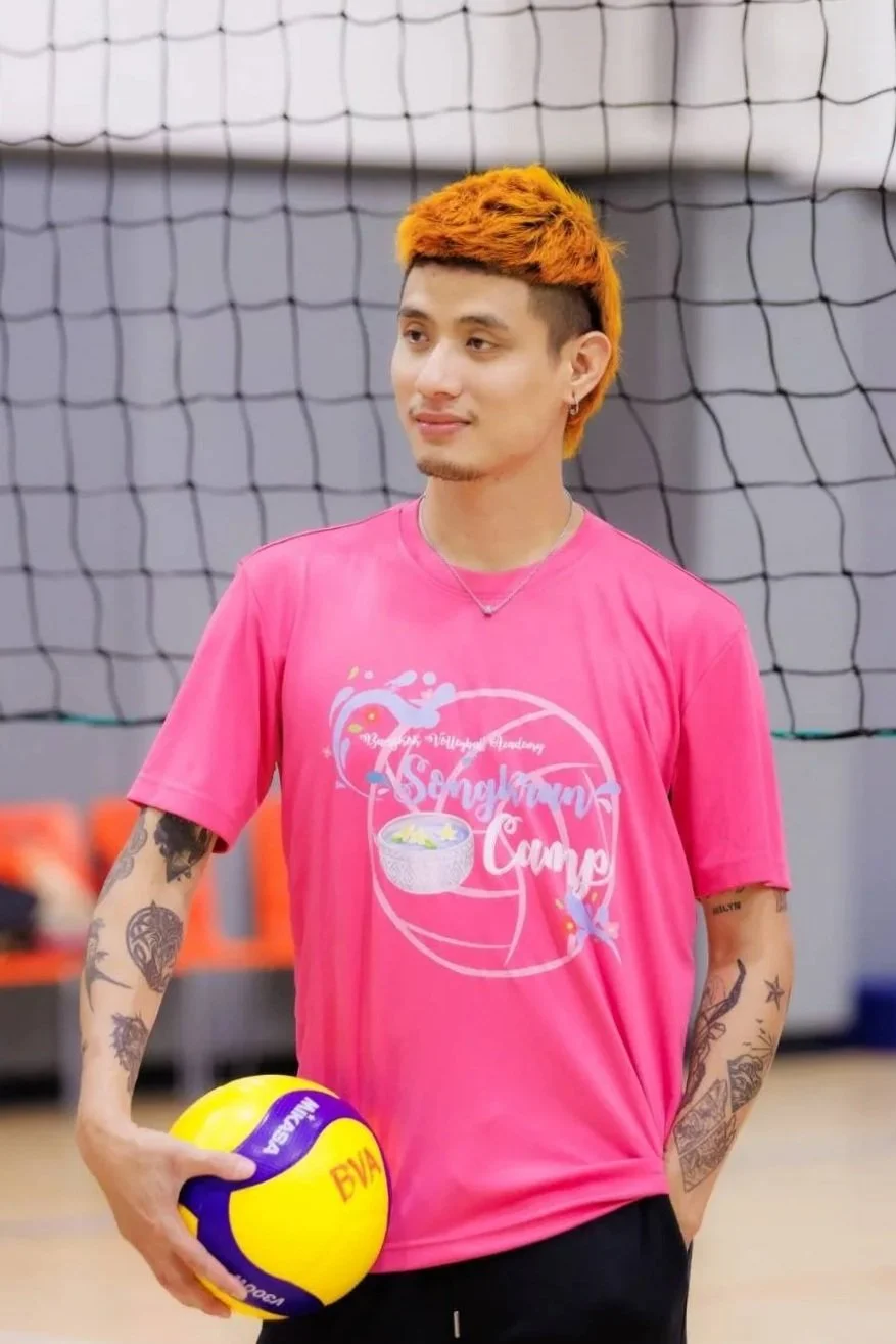 A young man with orange hair, tattoos on arms, wearing a pink T-shirt, holding a yellow and purple volleyball, standing in a gymnasium near a volleyball net.