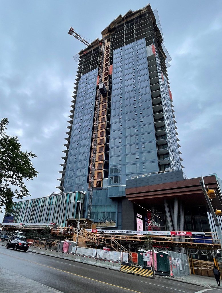 A tall high-rise building under construction with a crane on top, surrounded by construction barriers and equipment, with some finished glass windows and an overcast sky.