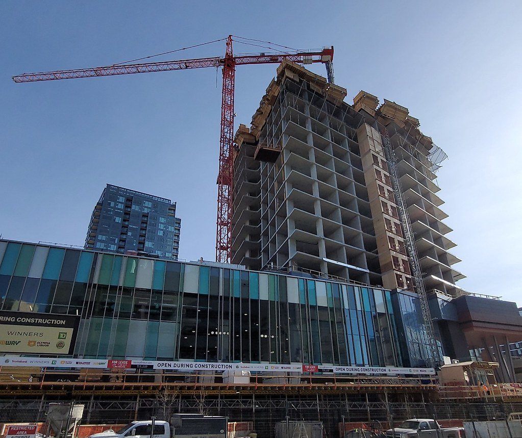 A high-rise building under construction with a red tower crane on top, glass windows on the lower floors, and construction banners on the fencing.