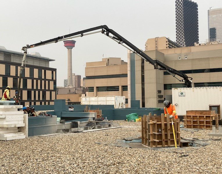 Construction workers on a rooftop using a concrete pump with a long arm to pour concrete into a form on a city rooftop. City buildings and a tower are visible in the background.