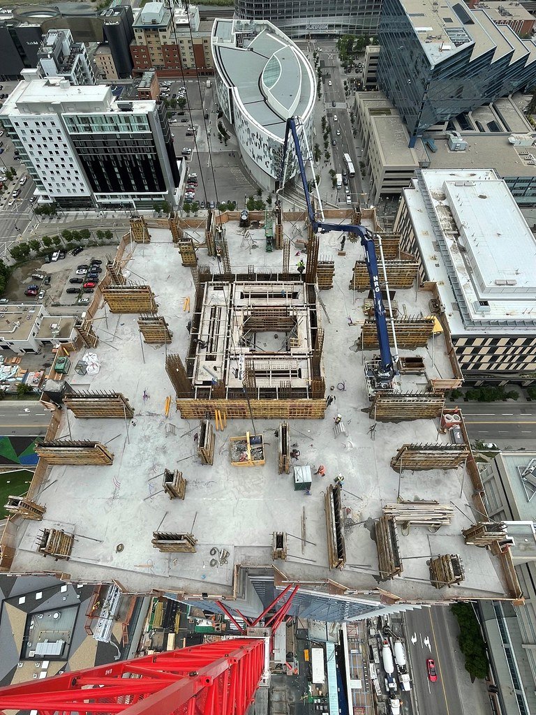 Construction site on the rooftop of a tall building in an urban area, with a crane lifting materials and workers present, surrounded by other high-rise buildings.
