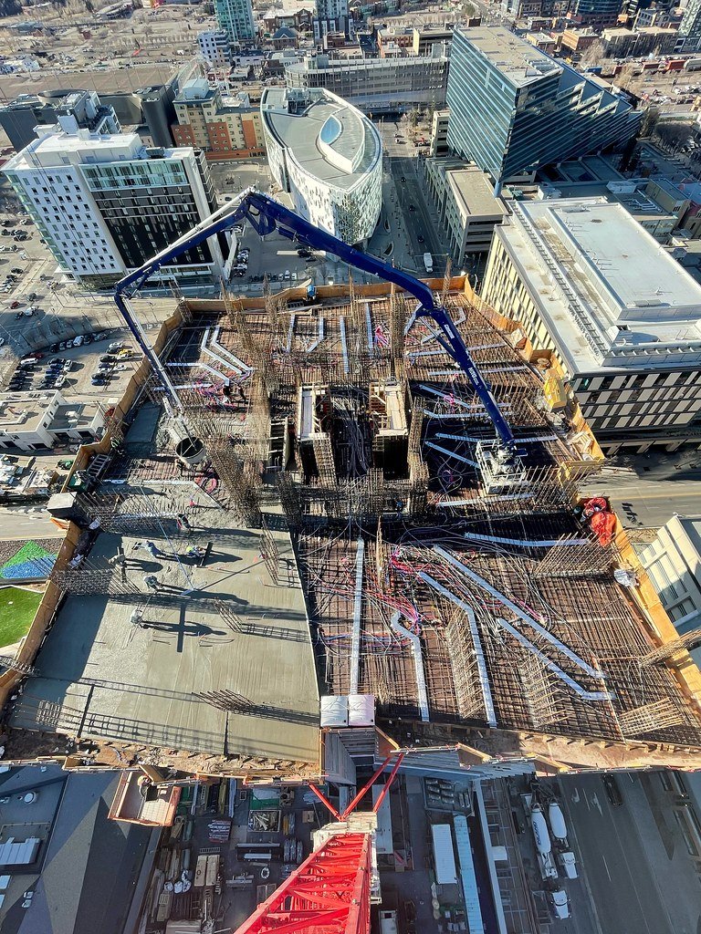 View from a high-rise building showing a rooftop construction site with scaffolding, rebar, and a concrete pump in an urban cityscape with multiple modern buildings surrounding it.