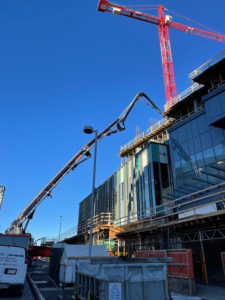 Construction site with a red tower crane, a concrete pump truck extending its arm, and a modern building under construction with glass windows and scaffolding.