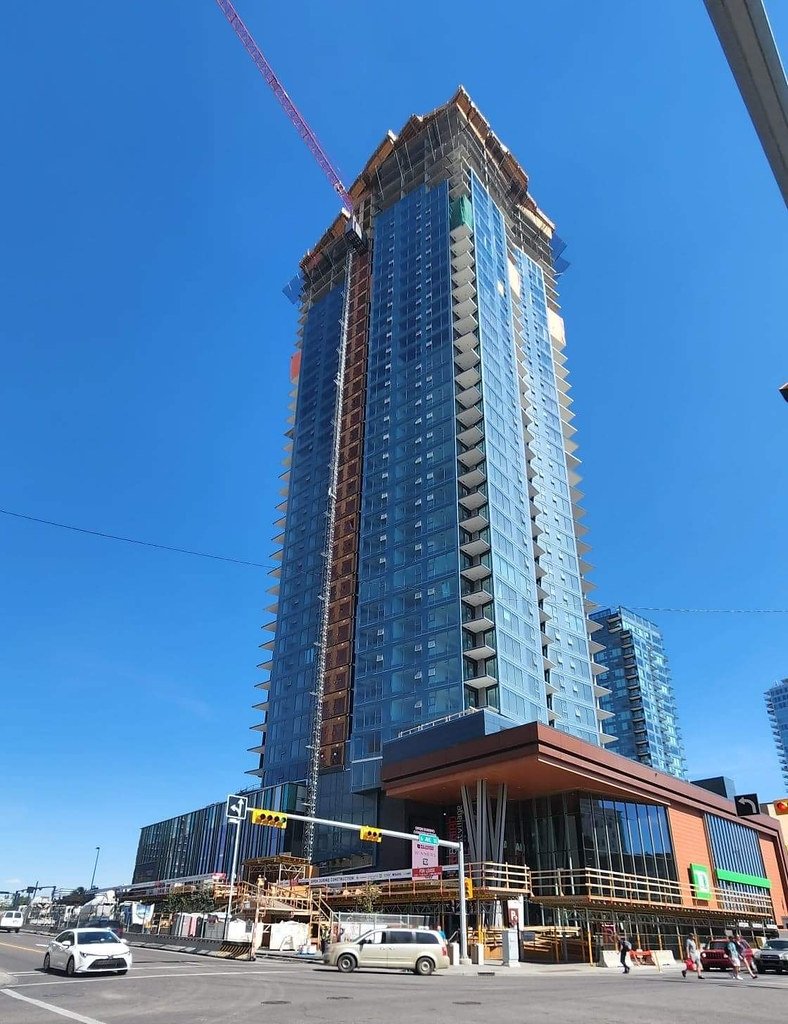 A tall building under construction with a purple crane on top, at an intersection with cars and pedestrians.