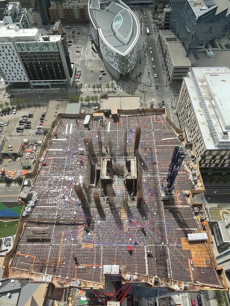 Construction site on the roof of a high-rise building, with steel rebar, wiring, and cranes, surrounded by city buildings and streets.