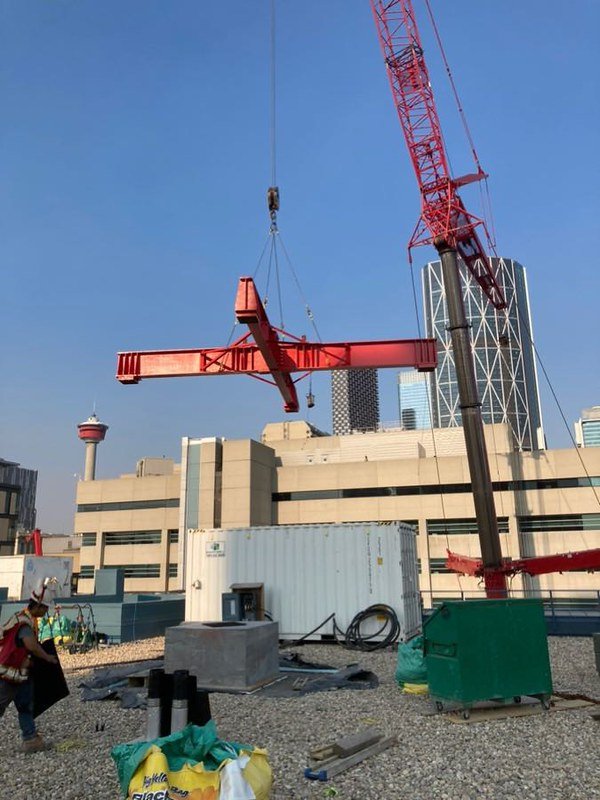 Construction crane lifting a red steel beam on a rooftop with city buildings and a tower in the background.