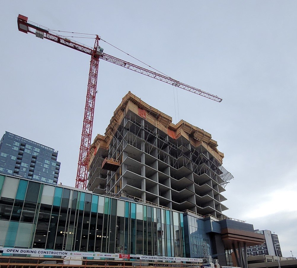 A tall building under construction with a large red crane on top, next to a modern glass building, and a cloudy sky in the background.