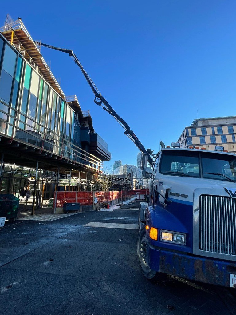 Construction site with a large truck and crane lifting materials above a modern glass building on a clear blue day.