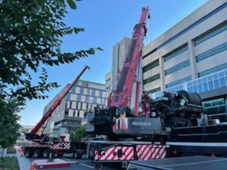 A large red mobile crane with extended boom is set up on a city street next to modern buildings, with a second smaller crane nearby, during daytime.