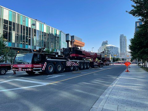 Large crane truck parked on city street next to modern glass buildings, with a city skyline in the background and clear blue sky.