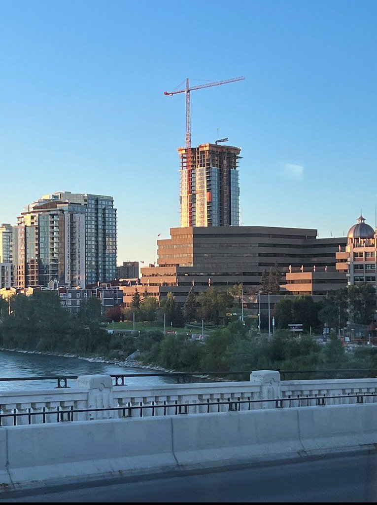 City skyline with high-rise buildings, including a building under construction with a crane, seen from a bridge over a river, during daylight with clear skies.
