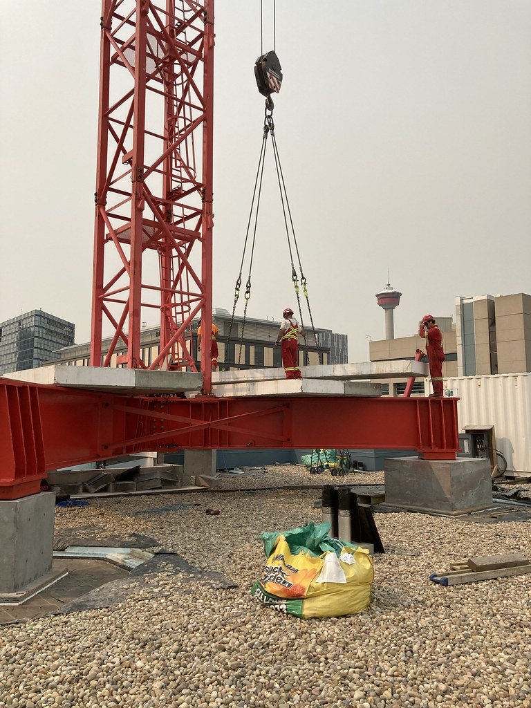 Construction workers in red safety gear working on a rooftop with a red crane, city buildings, and a tower in the background, on a gravel surface.