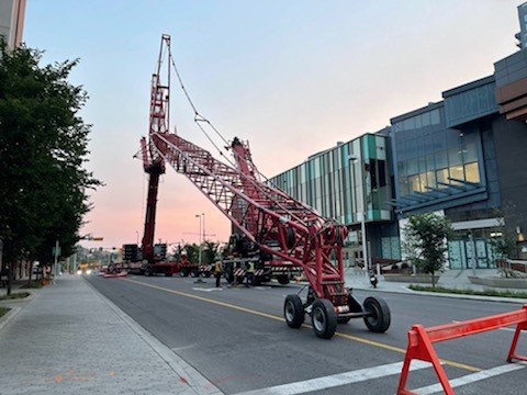 Large red construction crane positioned on a city street with modern buildings and trees in the background.