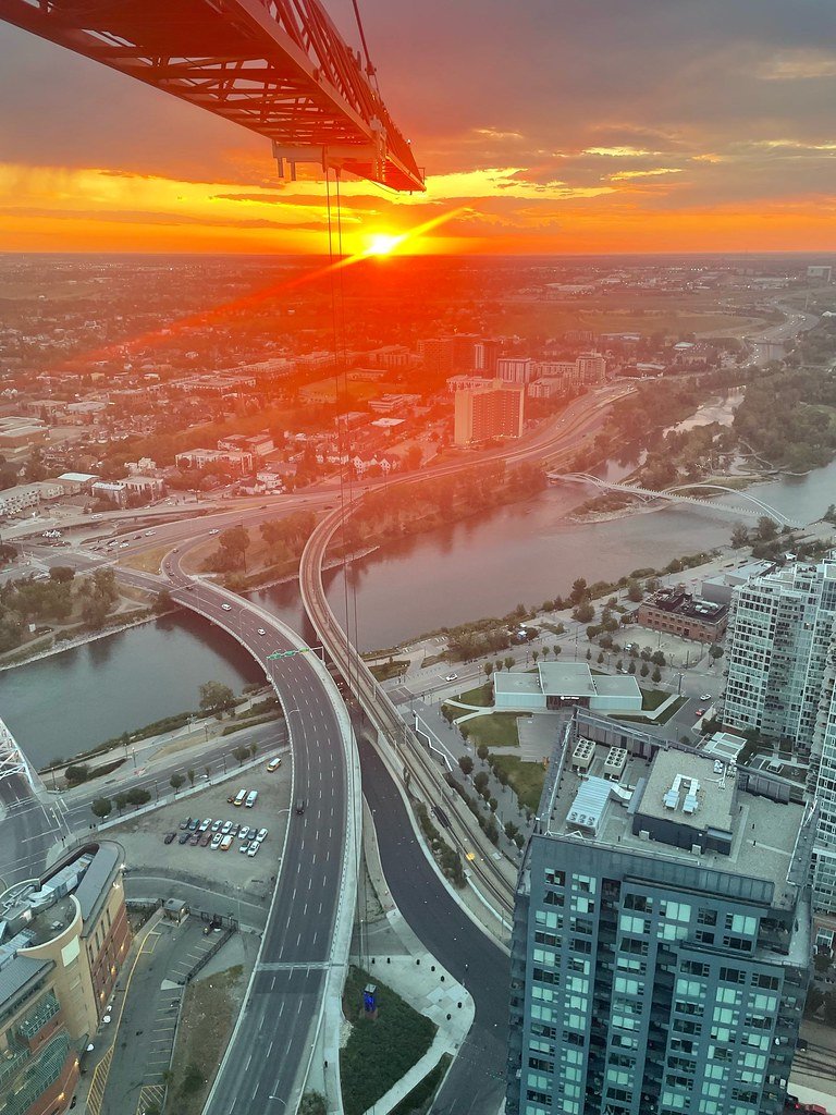Cityscape view from a tall building featuring a highway overpass, a river, and a sunset sky with clouds, with the building's edge and construction crane visible in the foreground.