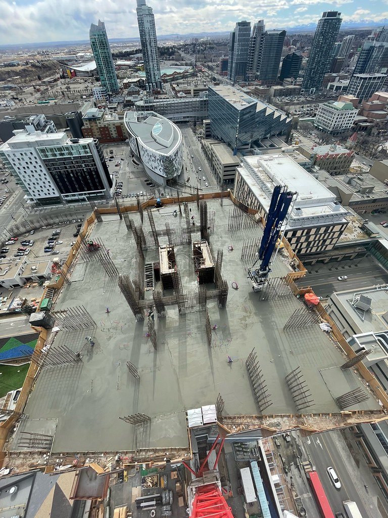 Looking down from a skyscraper construction site showing a construction area with concrete floor, steel rebar protruding, and a tower crane. Surrounding urban cityscape with modern high-rise buildings and streets below.