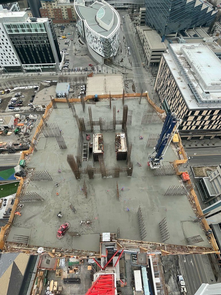 View from a high-rise building showing a construction site of a building with a concrete foundation, metal rebar structures, and a large blue crane on the right side. Surrounding the site are city streets and tall office buildings.