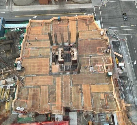 Top view of a building under construction with a wooden framework on a city street, with adjacent sidewalks, parked cars, and nearby buildings.