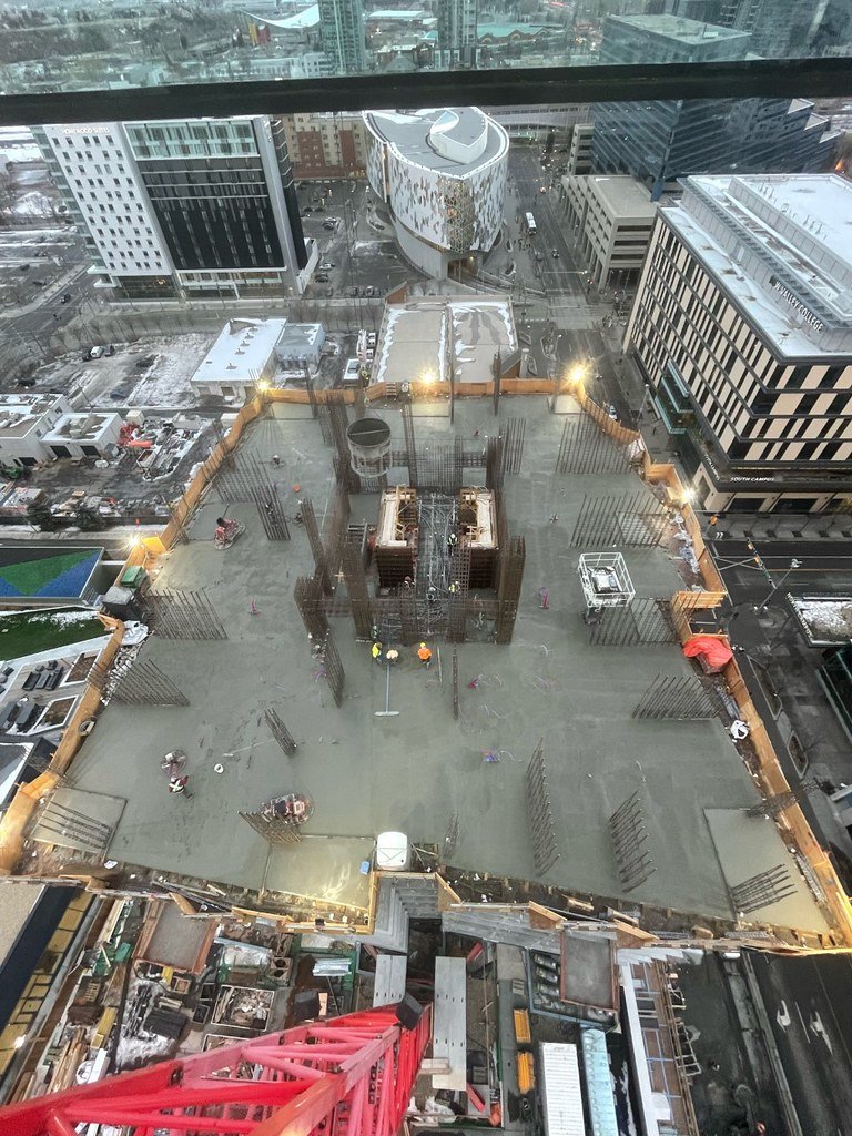 Bird's eye view of a construction site on a high-rise building with workers and concrete, surrounded by city buildings and streets.
