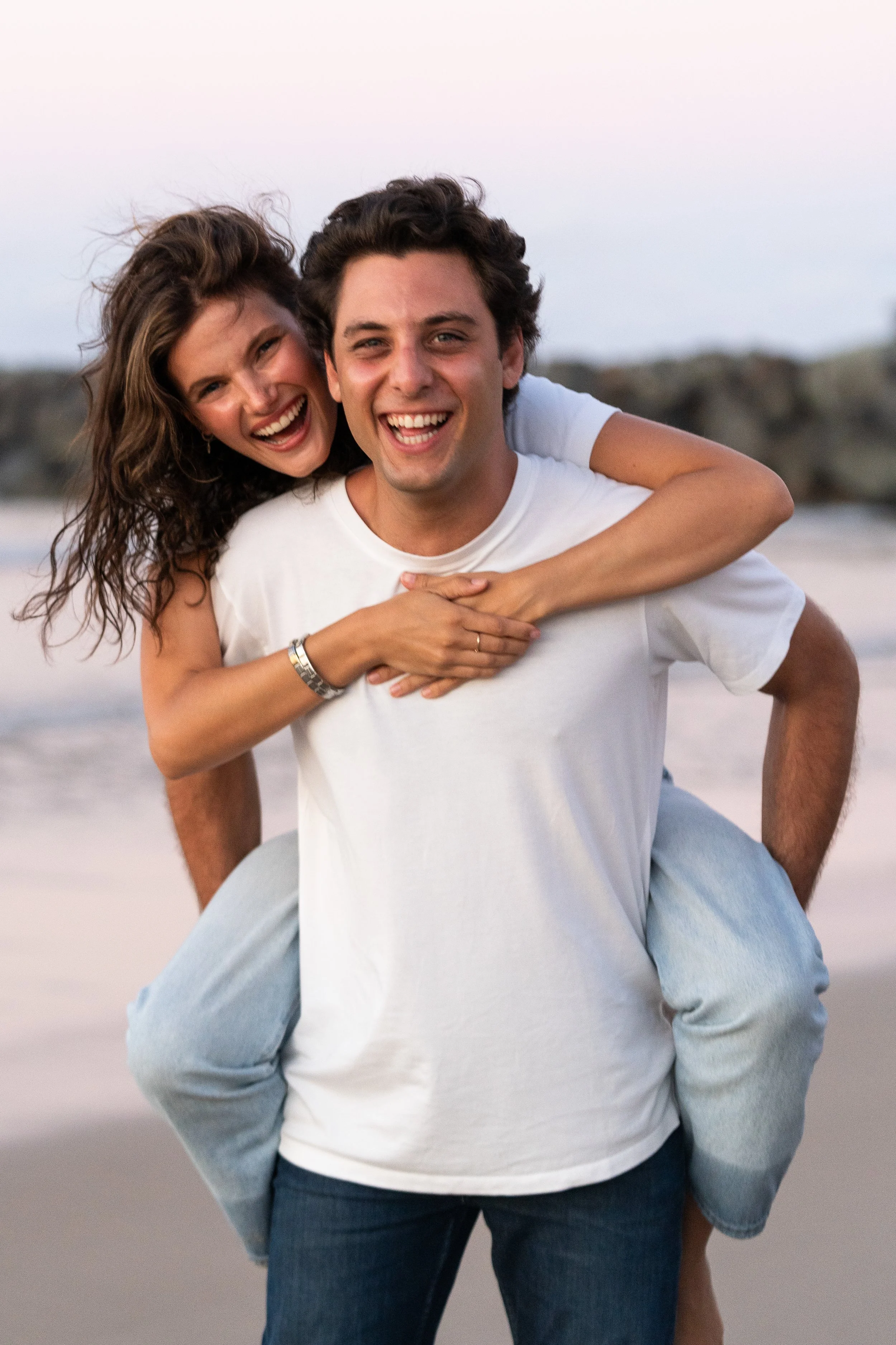 A young man giving a piggyback ride to a young woman on the beach, both smiling and looking happy during sunset.