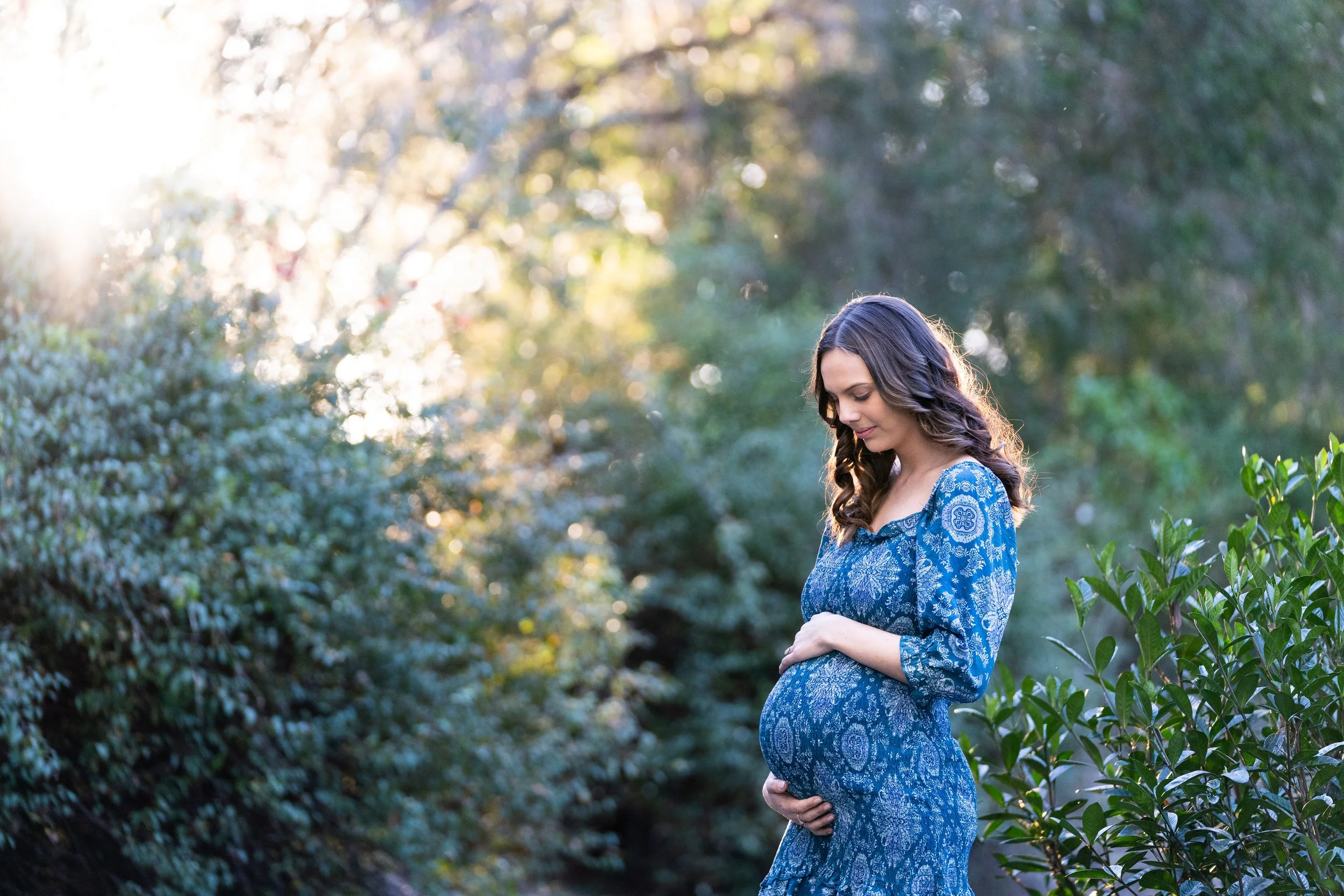 Pregnant woman in a blue patterned dress standing outdoors among green bushes and trees, walking in sunlight, gently holding her baby bump and looking down at it.