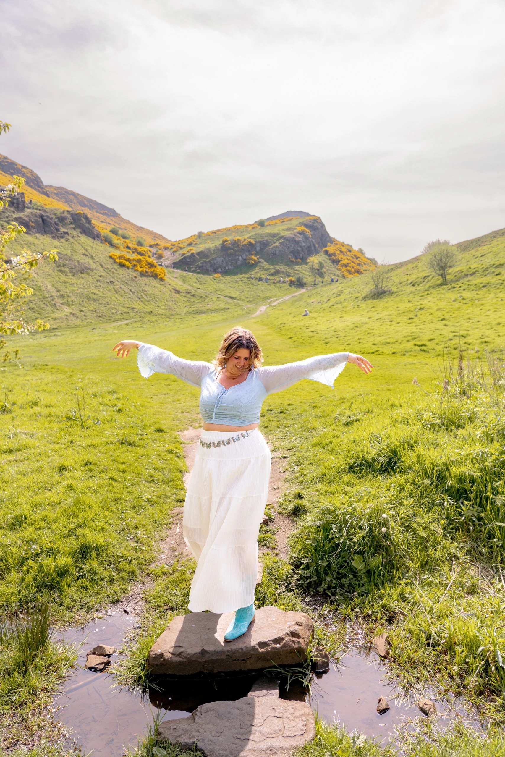 A woman standing on a rock in a stream with arms outstretched in a green meadow with rolling hills and yellow flowering bushes in the background.