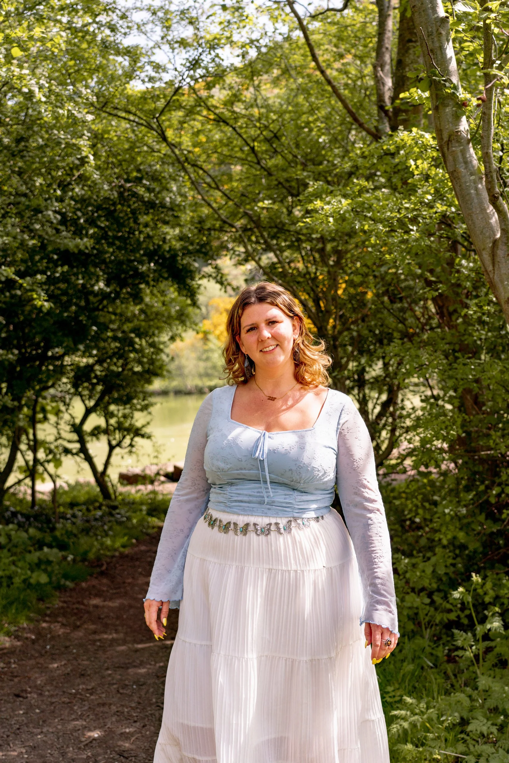 A woman with wavy blonde hair, wearing a light blue top and a long, flowing white skirt, standing on a wooded trail near a body of water during daytime.