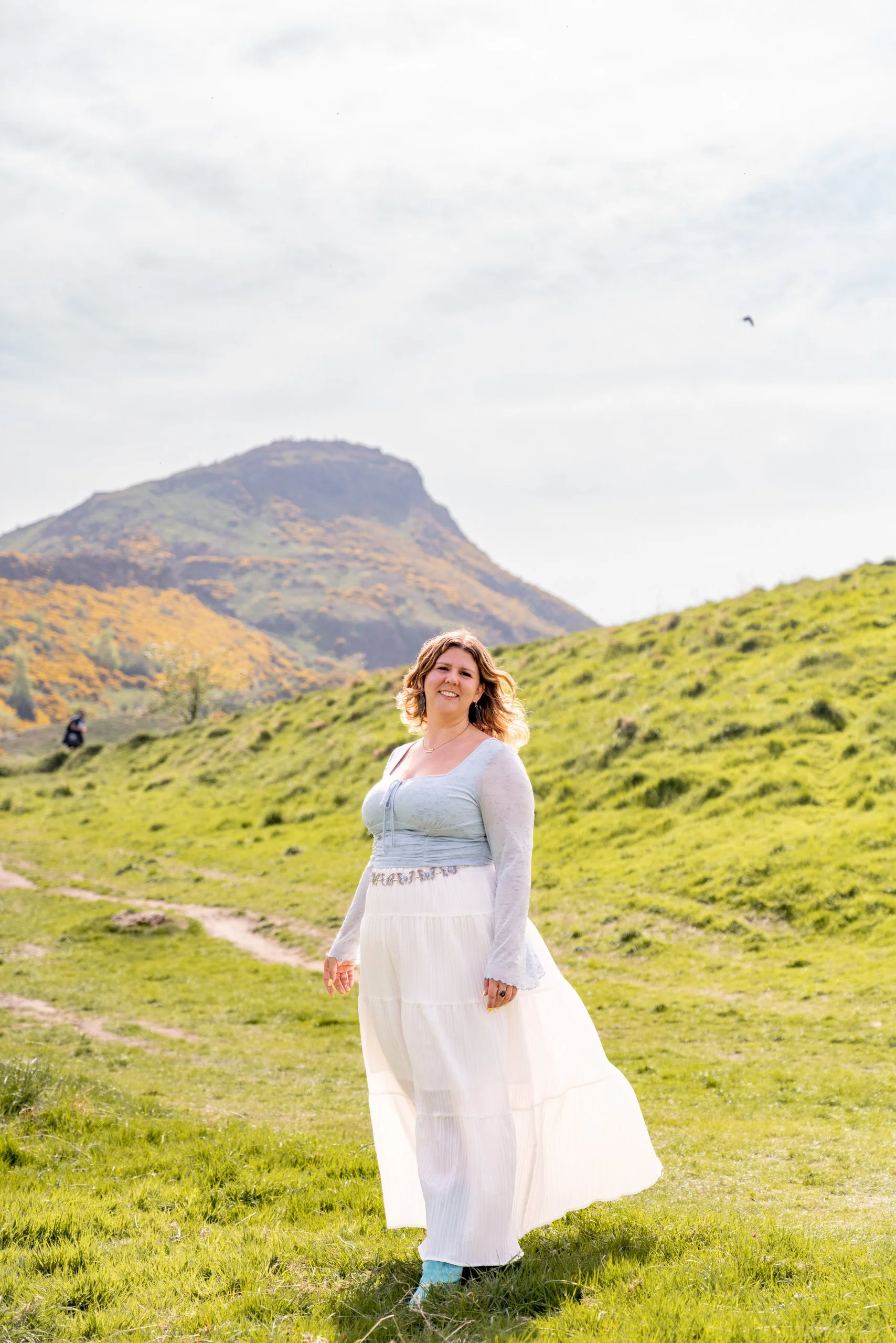 A woman in a long white skirt and light-colored top standing on a grassy hill with a mountain in the background and a cloudy sky overhead.