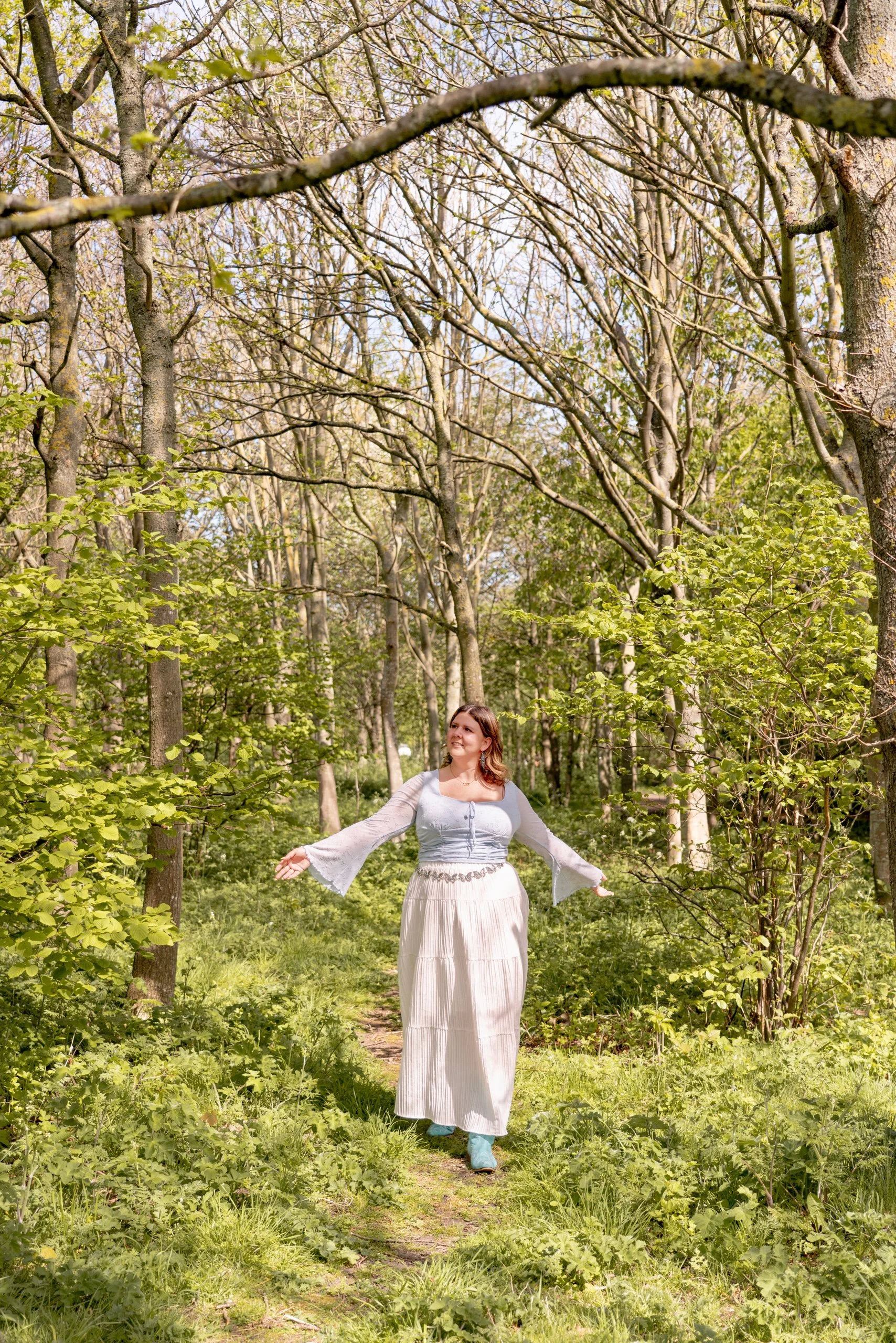 A woman in a long white skirt and blue top standing on a trail in a lush green forest with trees and bright sunlight.