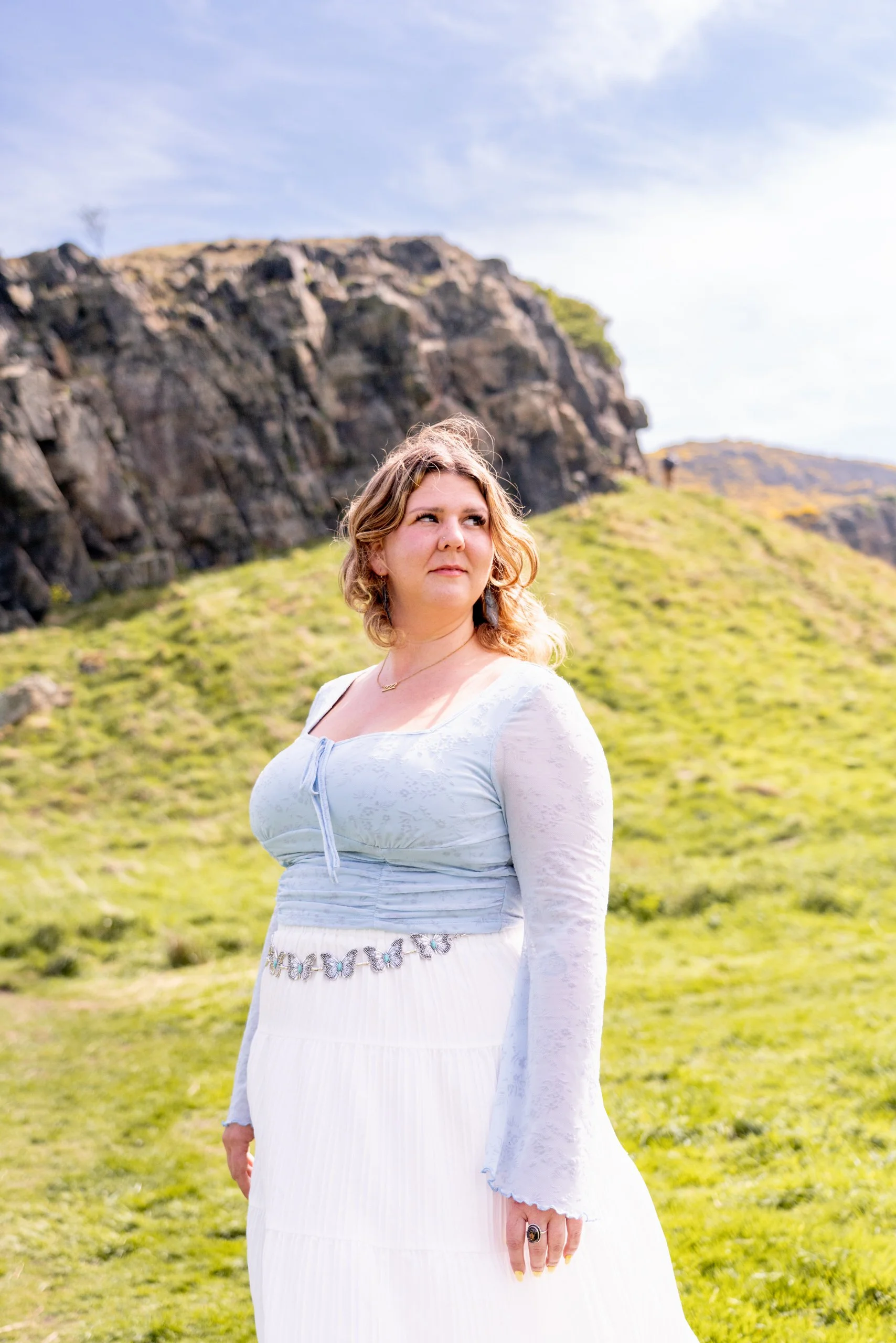 A woman with shoulder-length blonde hair wearing a white dress with a light blue bodice stands outdoors on a grassy hill with rocky cliffs in the background under a partly cloudy sky.