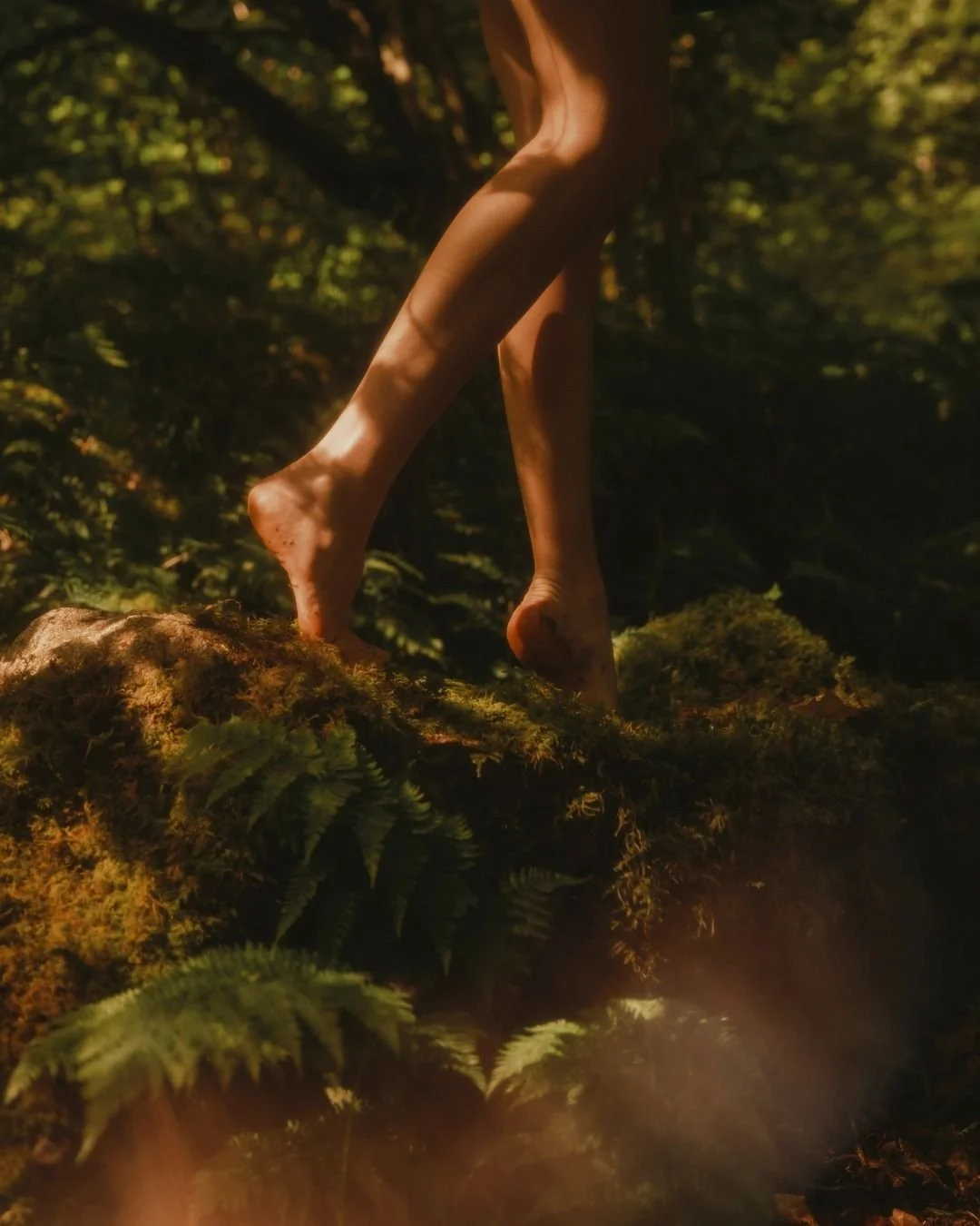 Close-up of human legs and feet standing on mossy ground in a forest, with sunlight filtering through trees.