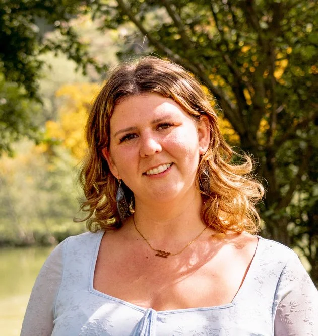A woman with shoulder-length wavy brown hair, smiling outdoors with trees and a body of water in the background.
