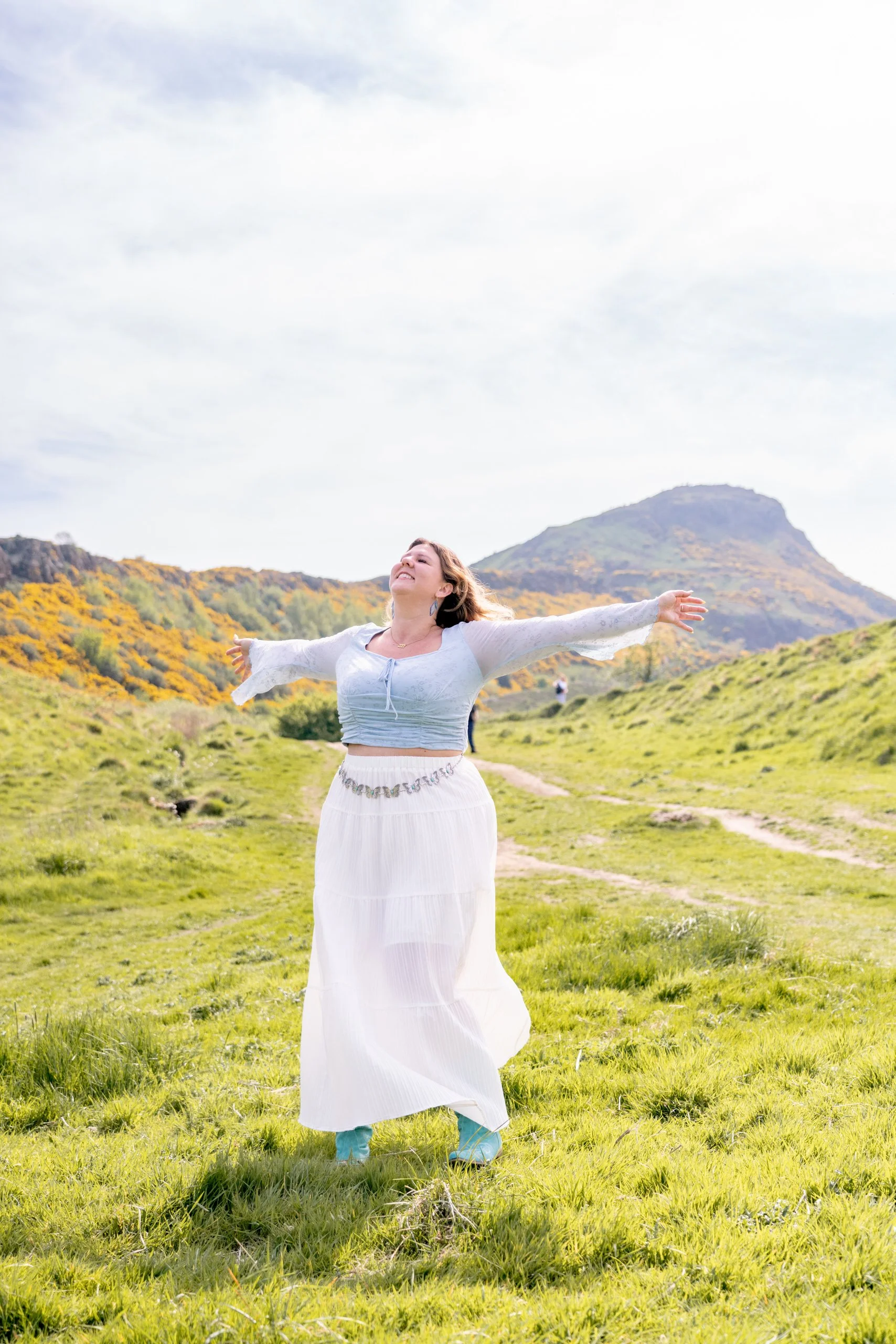 A woman standing in a green grassy field with her arms outstretched and her face tilted up, smiling. In the background, there are hills with yellow and green foliage under a partly cloudy sky.