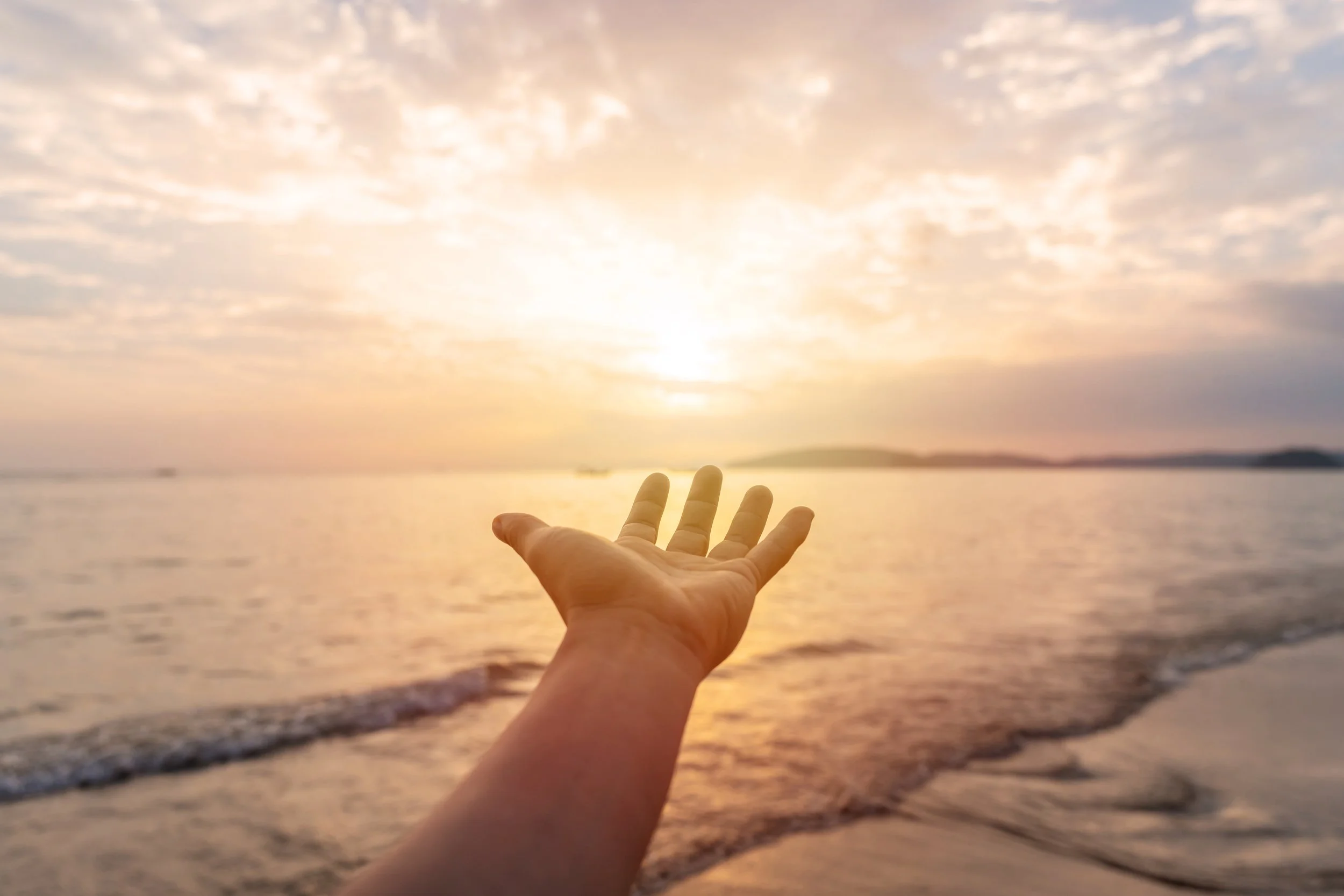 A person's hand reaching out towards a sunset over the ocean, with a partly cloudy sky and a distant landmass on the horizon.