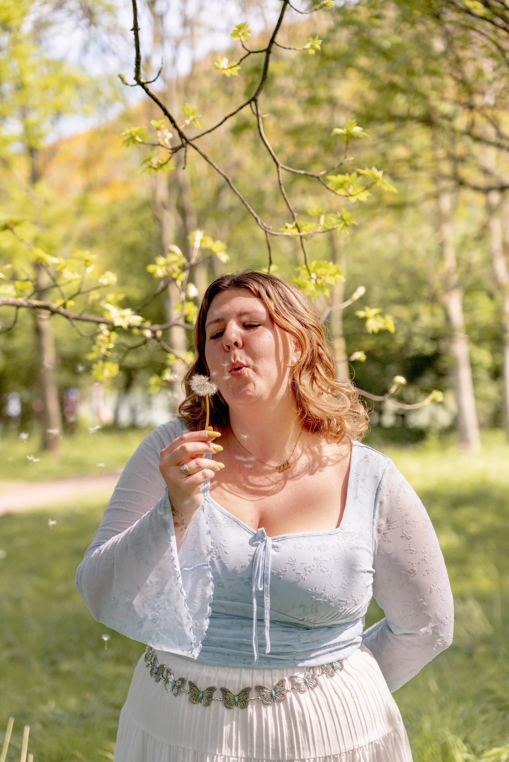 A woman with light brown hair wearing a white blouse and skirt, standing outdoors under green trees, blowing on a dandelion.