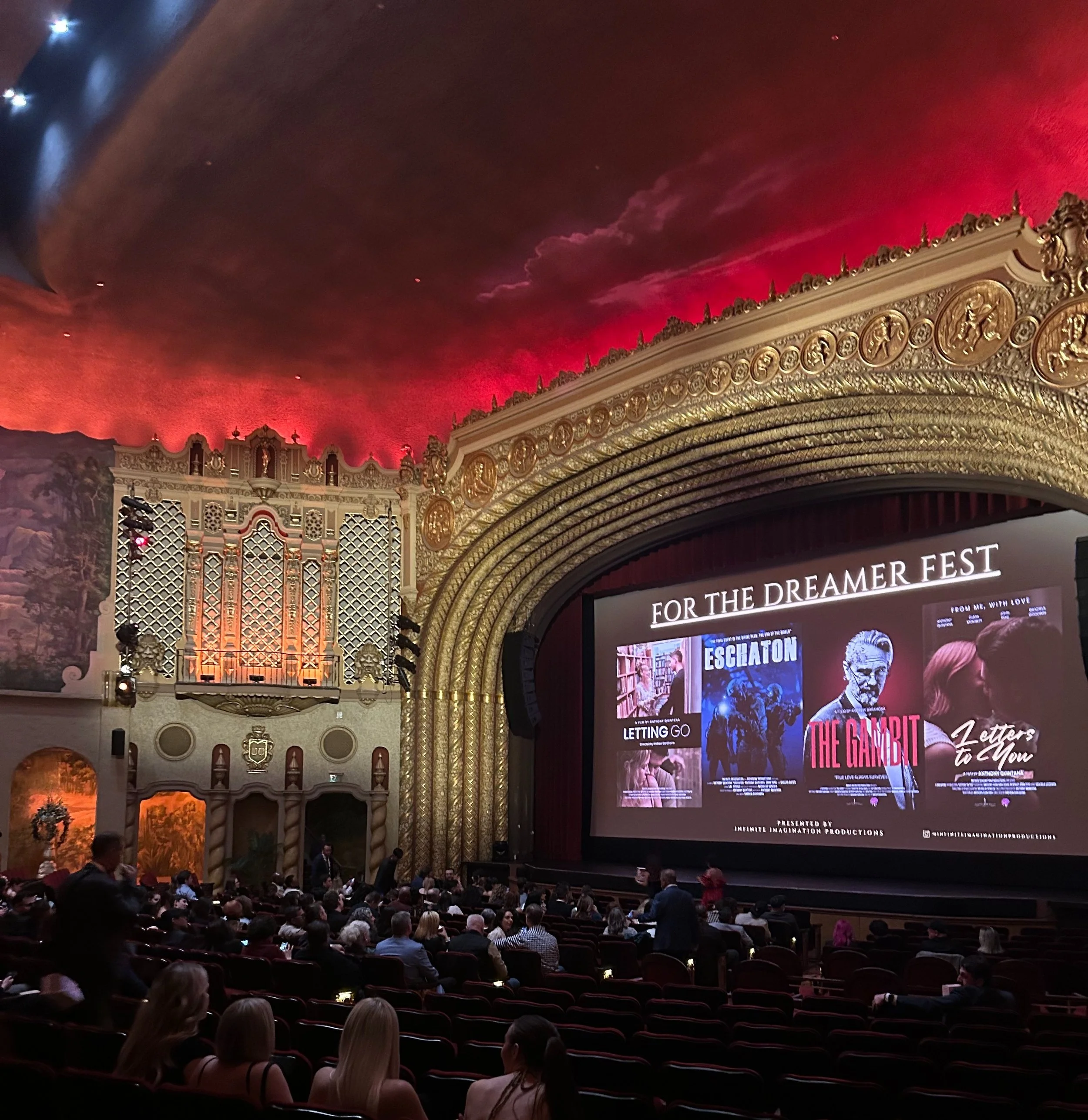 Interior of a vintage movie theater with a large audience, ornate gold and white decorations, a painted ceiling with red and dark clouds, and a screen displaying posters for films titled Letting Go, Escatron, The Gambit, and Letters to You.