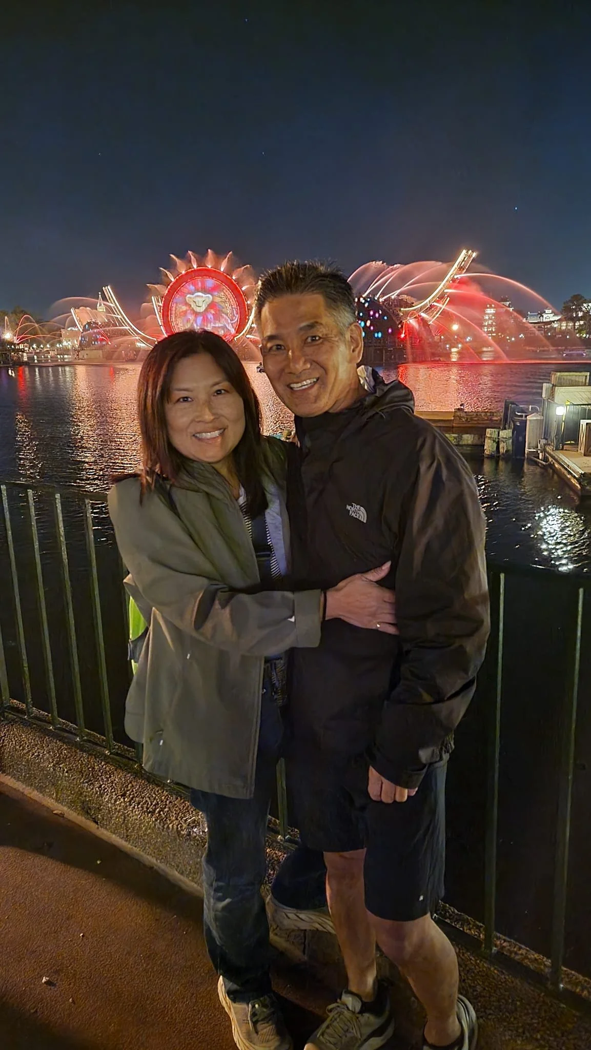 A couple smiling and embracing in front of a night fountain display by a body of water.